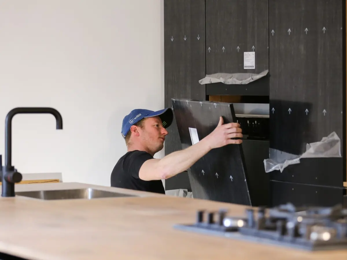 Monteur schuift een oven op zijn plaats in een zwarte inbouwkast, op de voorgrond een aanrecht met kraan en kookplaat